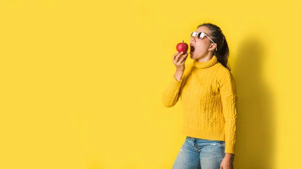 Cheerful young woman on yellow background in studio with a red apple in her hand she is opening her mouth to eat apples. The concept good health.