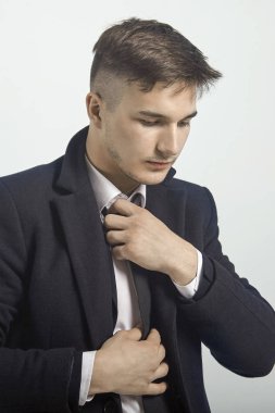 Siyah kravatını düzelten yakışıklı bir adamın portresi. Klasik siyah takım elbiseli, beyaz gömlekli, resmi tarzda, ciddi görünümlü. Işık arka plan, stüdyo, izole edilmiş, kopyalama alanı. Moda saç stili, küçük sakal..