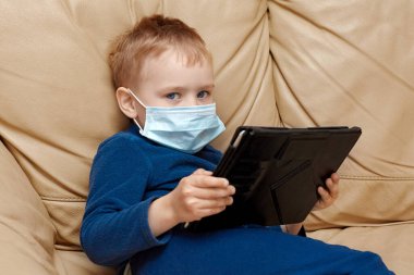 Elinde tablet kâğıdı olan, tıbbi maske takan sevimli erkek bebek deri koltukta oturuyor. Mavi pijama giyiyor, duygusal yüz ifadesi takınıyor. Hastalık veya virüs konsepti, hastane veya karantina, kopyalama alanı.
