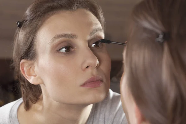 Young attractive woman does make up with mascara brush, looking into reflection in mirror. Beautiful female with dark hair and natural beauty. Warm background, soft everyday look. Copy space, indoors.