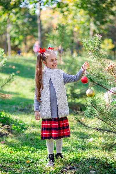 Portrait of a little girl near the Christmas tree. Girl decorates a Christmas tree in the forest with Christmas balls. — Stock Photo, Image