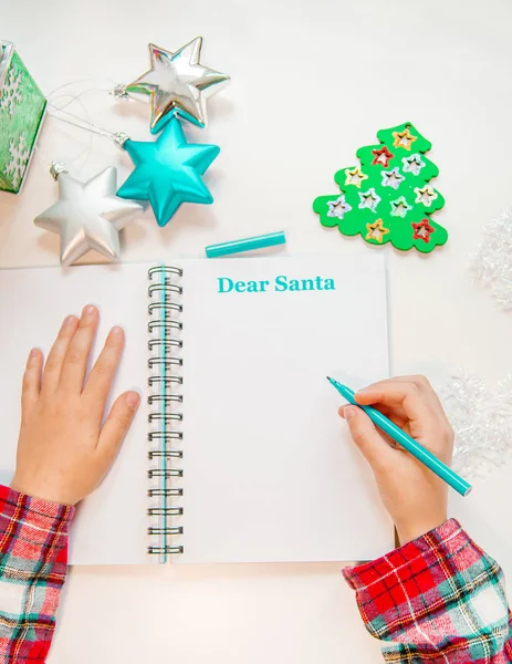 Querida carta de Pai Natal, cartão de Natal. Uma criança segurando uma caneta escreve em uma folha branca em um fundo de madeira com decoração de Ano Novo . — Fotografia de Stock