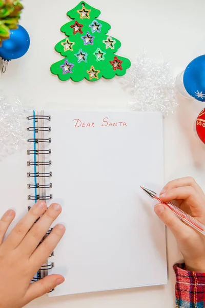 Dear Santa letter, Christmas card. A child holding a pen writes on a white sheet on a wooden background with New Year\'s decor.