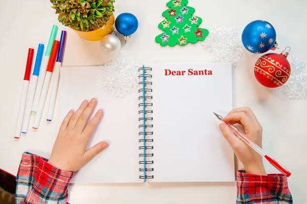 Dear Santa letter, Christmas card. A child holding a pen writes on a white sheet on a wooden background with New Year's decor.