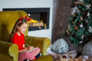 Noel Baba şapkalı tatlı bir kız Noel ağacının yanında Noel Baba 'ya mektup yazıyor. Mutlu çocukluk, arzularını tatmin etme zamanı..