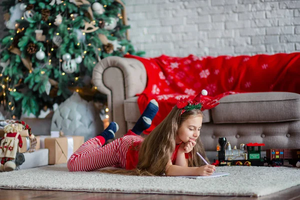 Une jolie fille dans un chapeau de Père Noël écrit une lettre au Père Noël près du sapin de Noël. Bonne enfance, un temps pour satisfaire les désirs . — Photo