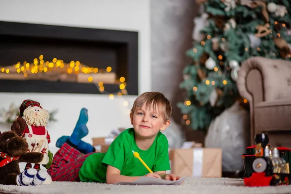 Joyeux garçon est une lettre au Père Noël, près du sapin de Noël. Bonne enfance, temps pour réaliser les vœux . — Photo