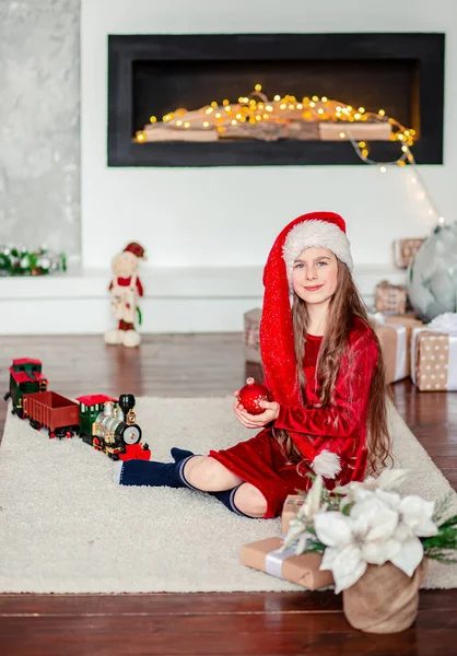 Portrait d'une jolie fille souriante dans un chapeau de Père Noël avec une boule rouge dans les mains, assise près d'un arbre de Noël décoré . — Photo