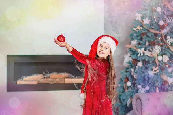 Portrait of a cheerful cute smiling girl in a santa hat with a red ball in hands, sitting near a decorated Christmas tree. — Stock Photo, Image