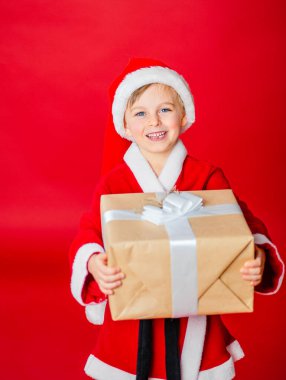 Kırmızı arka planda Noel Baba kıyafeti giymiş neşeli bir çocuk elinde bir hediye tutuyor..