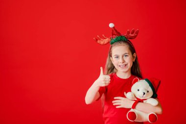 Kafasında Noel pijaması ve boynuz bandajı olan neşeli bir kızın portresi. Kız oyuncak ayıya sarılıyor..
