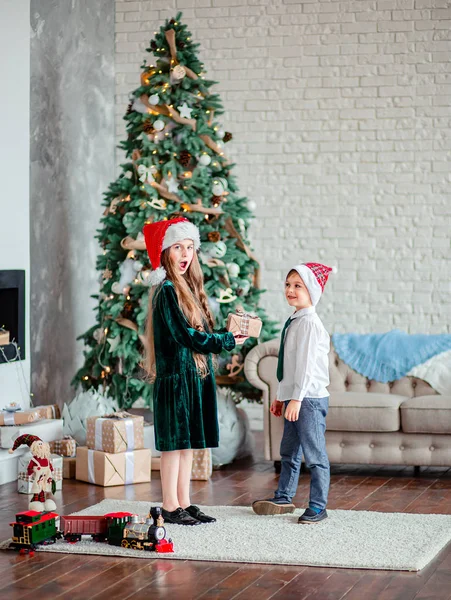 Frère et sœur donnent et déballer des cadeaux sous le sapin de Noël, assis près de la cheminée, le matin de Noël . — Photo