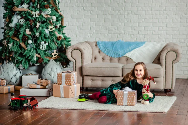 Petite fille sous le sapin de Noël dans le chapeau du Père Noël avec des cadeaux sous le sapin de Noël près de la cheminée . — Photo