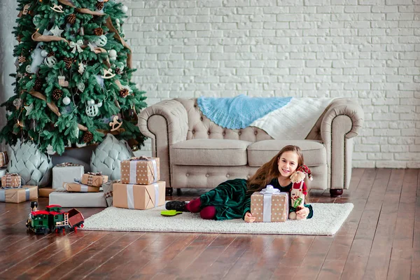 Petite fille sous le sapin de Noël dans le chapeau du Père Noël avec des cadeaux sous le sapin de Noël près de la cheminée . — Photo