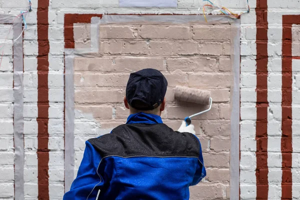 A painter in a black and blue robe paints a white wall in pink.