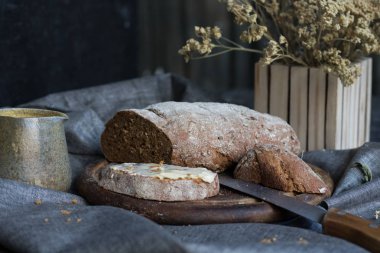 Taze ekmek, ahşap tahtada tereyağlı lezzetli bir dilim ve bir bardak içecek, bıçak ve çizgili arka planda bulanık çiçek saksısında kurutulmuş çiçek.