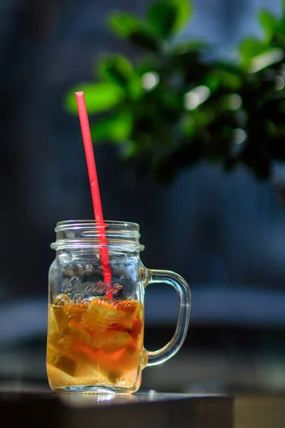 glass of drink with ice, straw and fruit on wooden table and blurred twig with green foliage