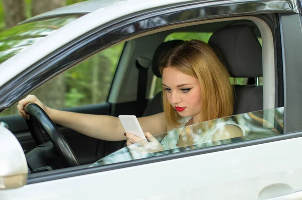 Hermosa chica detrás del volante de un coche, buscando teléfono — Foto de Stock