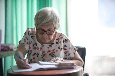 Asyalı yaşlı bir kadın cam kenarında oturuyor ve masaya yazı yazıyor.