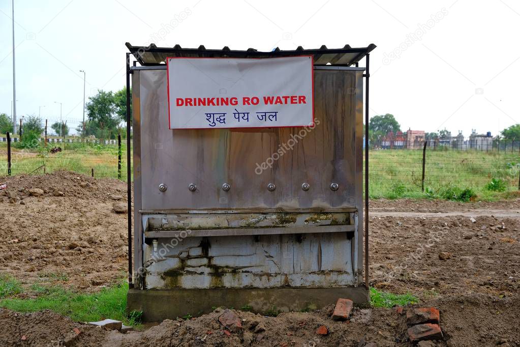 A metal structure in the middle of mud trenches with a sign in English and Hindi about the availability of drinking water.