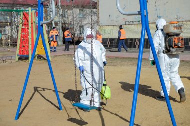 RAMENSKOYE, RUSSIA - 26 Mart 2020: Koruyucu giysili ve maskeli kadın çocuk oyun alanını dezenfekte ediyor. COVID-19 enfeksiyonu ve kitle alımının yayılmasını önlemek için önlemler