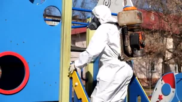 Un uomo in tuta protettiva e maschera disinfetta un parco giochi per bambini. Misure per prevenire la diffusione dell'infezione da COVID-19 e l'assunzione di massa — Video Stock