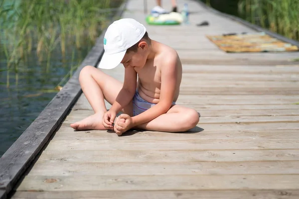 Il ragazzo, seduto su un pavimento di legno, con la mano tira fuori una scheggia nel piede . — Foto Stock