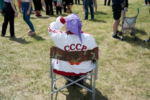 クラズノヤルスク州シャリーポフスキー地区パルナイア 7月2019 碑文を持つトラックスーツの高齢者女性ソ連はカラタグ音楽祭の人々を背景に清掃中の折りたたみ椅子に座っています — ストック写真