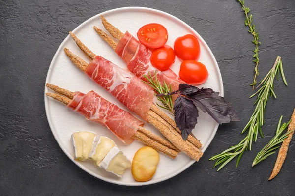 Traditional Italian food is grissini bread with prosciutto, cheese and tomatoes with herbs on a plate on a dark background. Healthy style. Copy space — Stock Photo, Image