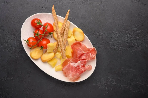 Traditional Italian food is grissini bread with prosciutto, cheese and tomatoes with herbs on a plate on a dark background. Healthy style. Copy space — Stock Photo, Image