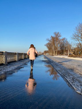Pembe ceketli bir kadın açık bir günde setin kenarında yürüyor ve su birikintilerine yansıyor.