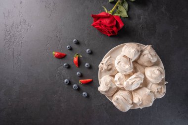 Homemade popular dessert meringue on a white plate on a black background. Nearby are strawberry and blueberry berries and red rose flower