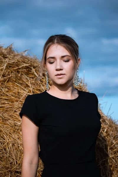 Style fashion on street. A girl with bundle hair in a earrings, black dress and white sneakers. Female portrait. Model posing. A girl in a dress on a haystack background. Fashion photography