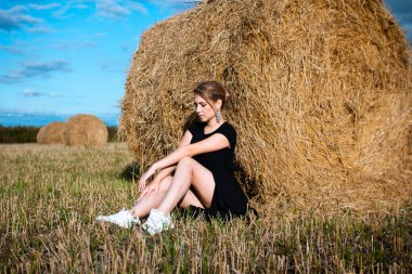 Sokakta moda. Küpeli, siyah elbiseli ve beyaz spor ayakkabılı bir kız. Kadın portresi. Model pozu. Samanlık arka planda elbise giymiş bir kız. Moda fotoğrafçılığı