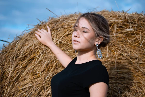 Style fashion on street. A girl with bundle hair in a earrings, black dress and white sneakers. Female portrait. Model posing. A girl in a dress on a haystack background. Fashion photography