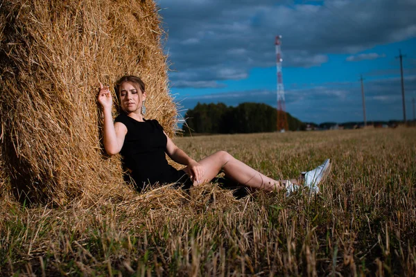 Style fashion on street. A girl with bundle hair in a earrings, black dress and white sneakers. Female portrait. Model posing. A girl in a dress on a haystack background. Fashion photography