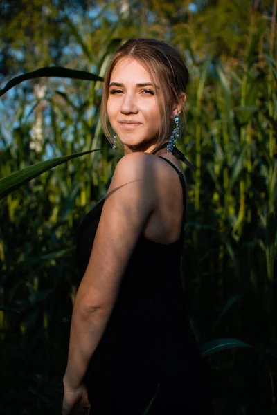 Close-up of young woman in black dress, necklace and earrings between green leaves in a corn field in summer. Jewelry fashion style photo session.