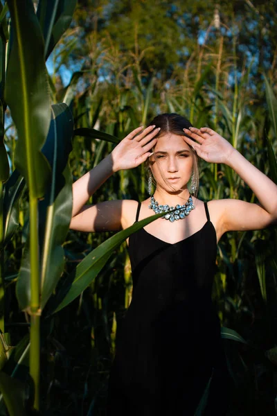 Close-up of young woman in black dress, necklace and earrings between green leaves in a corn field in summer. Jewelry fashion style photo session.