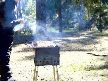 Yanan kömürün yanında ızgara yapmaya hazırlanan adam. Arka planda orman var. Barbekü ızgarası, barbekü.. 