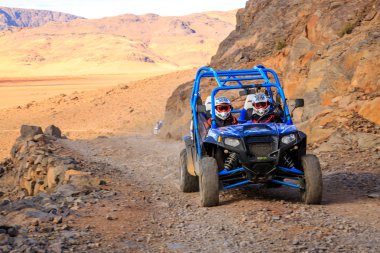 Merzouga, Morocco - Feb 21, 2016: Blue Polaris RZR 800 and pilots crossing on a mountain road in Morocco desert near Merzouga. Merzouga is a small village located in the Saharan south-eastern Morocco. clipart