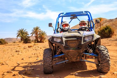 Merzouga, Morocco - Feb 26, 2016: front view on blue Polaris RZR 800 with it's pilots in Morocco desert near Merzouga. Merzouga is famous for its dunes, the highest in Morocco. There is a palm grove i clipart