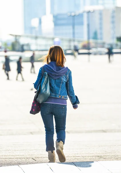 La defense, Francia- 10 de abril de 2014: vista trasera de una mujer morena caminando por una calle de centro de negocios. Lleva vaqueros azules y una bolsa. — Foto de Stock