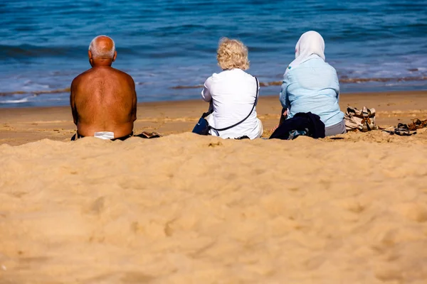 Saint-Jean de Luze, Francia - 28 de septiembre de 2016: familia sentada en toallas de playa en la arena. Tres personas, un hombre y dos mujeres. Llevan ropa de ciudad y un sombrero en la cabeza para protegerse de — Foto de Stock