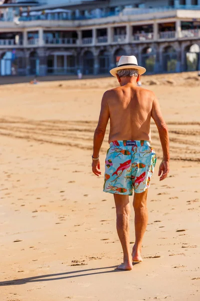 Saint-Jean de Luze, Francia - 28 de septiembre de 2016: Hombre mayor caminando en una playa al borde del agua. Está en topless en la playa y lleva pantalones cortos de flores. Hay hoteles en el fondo — Foto de Stock