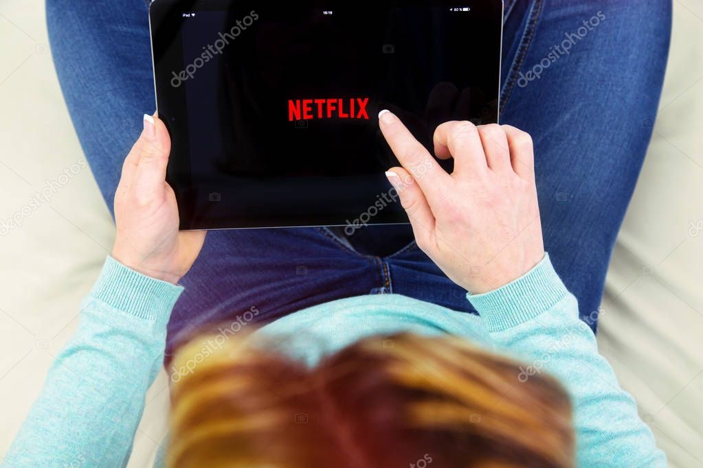 Benon, France - January 21, 2018: woman sitting cross-legged on her couch and using her touch pad to connect to the Netflix app. Netflix Inc. is an American multinational entertainment company founded on 1997