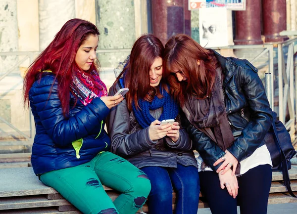 Amigos adolescentes usando teléfonos inteligentes en el banco en la plaza —  Fotos de Stock