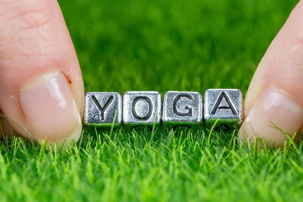 Mot YOGA écrit en lettres métalliques posées sur l'herbe et tenu entre les doigts d'une femme. Concept de bien-être, de soins du corps — Photo
