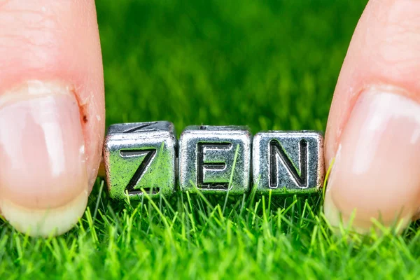 Слово ZEN написане металевими літерами, покладеними на траву і тримається між пальцями жінки. Концепція оздоровчого фону — стокове фото