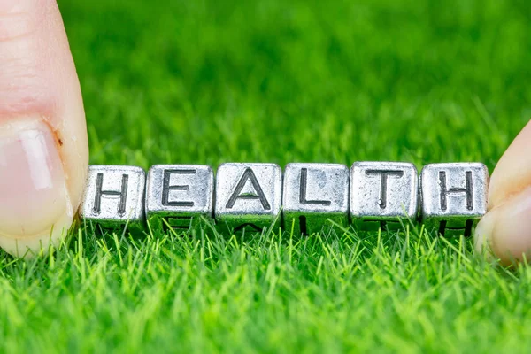 Word Gezondheid geschreven in metalen letters op gras gelegd en tussen de vingers van een vrouw gehouden. Concept van gezondheidsachtergrond — Stockfoto