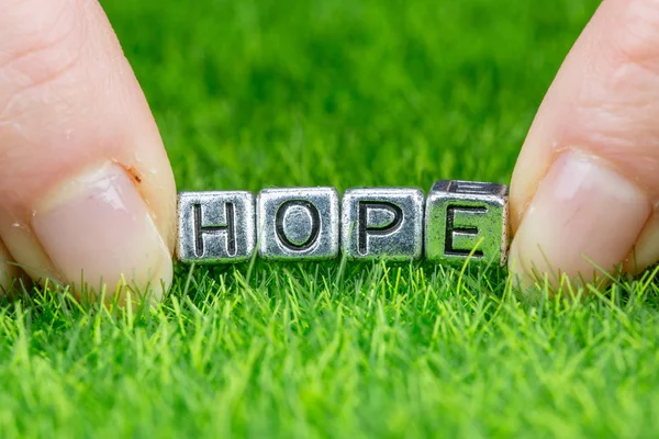 Слово HOPE написане металевими літерами, покладеними на траву і тримається між пальцями жінки. Концепція прагнення, фон надії — стокове фото